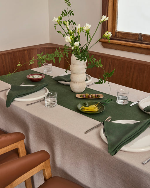 A dining table set for two features an Olive 100% French Flax Linen Table Runner by Bed Threads, a beige tablecloth, green napkins, white plates, clear glasses, cutlery, a ceramic vase with white flowers, and small dishes of olives and pickles.