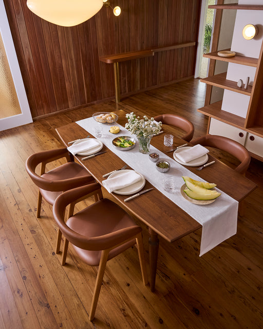 A wooden dining table set for four with tan chairs, white plates, glassware, food, and flowers in a vase rests on the Bed Threads White 100% French Flax Linen Table Runner in a wood-paneled room with wood floors.