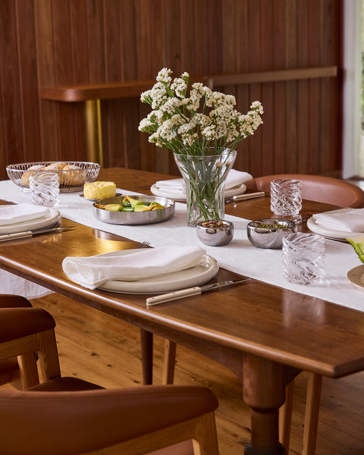 A wooden dining table set for a meal features the Bed Threads White 100% French Flax Linen Table Runner, white napkins, glassware, plates of food, and a vase of white flowers; wooden walls and chairs add to the cozy atmosphere.