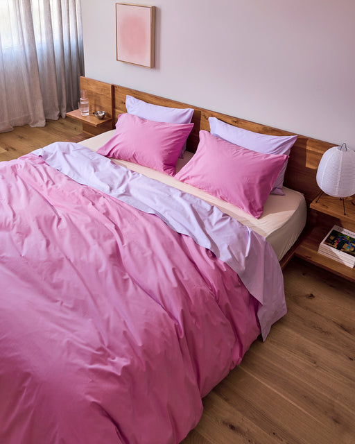 A bed featuring the Bed Threads Violet 100% Organic Cotton Flat Sheet, two pillows, and a wooden headboard. A lamp and glass of water rest on bedside tables as daylight filters through sheer curtains.