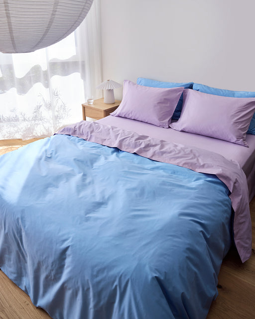 A neatly made bed features the Bed Threads Sky & Violet 100% Organic Cotton Bedding Bundle in light purple and blue. Sunlight filters through sheer white curtains onto a wooden bedside table with a small lamp.