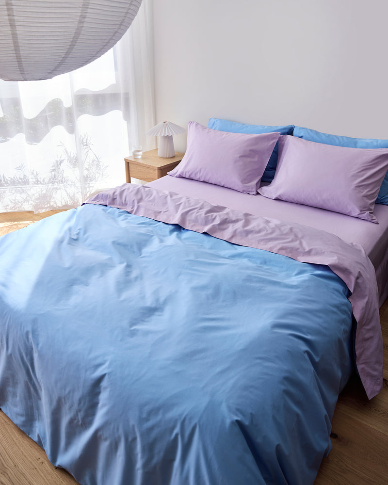 A neatly made bed features the Bed Threads Sky & Violet 100% Organic Cotton Bedding Bundle in light purple and blue. Sunlight filters through sheer white curtains onto a wooden bedside table with a small lamp.