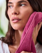 A woman with long brown hair gently presses a Berry 100% Cotton Terry Face Washer by Bed Threads to her cheek. Calm and wearing a white top, she looks off to the side, showcasing this soft, absorbent set of two face washers. - view