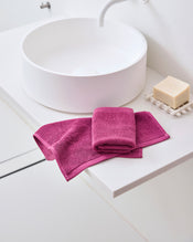 A modern white bathroom sink features Bed Threads Berry 100% Cotton Terry Face Washers (Set of Two) and a magenta towel neatly on the counter. A bar of soap on a dish beside the sink completes the clean, minimalistic look. - view