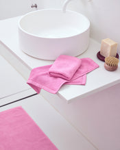 A modern white bathroom sink with a bar of soap, a brush, and two Bubblegum 100% Cotton Terry Face Washers by Bed Threads folded on the countertop. A matching pink bathmat completes the minimal, tidy scene designed to be gentle on skin. - view