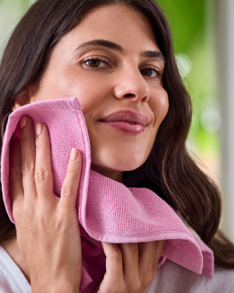A woman with long brown hair smiles gently while holding a Bubblegum 100% Cotton Terry Face Washer (Bed Threads, set of two) to her cheek. The soft, absorbent fabric looks gentle on skin. The softly blurred background features green and neutral tones.