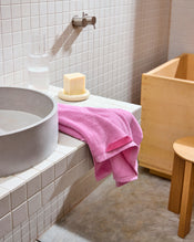 A modern bathroom with a white tiled sink, round gray basin, and a Bed Threads Bubblegum 100% Cotton Terry Hand Towel draped over the counter, with a glass of water, soap dish, wooden stool, and tub in the background. - view