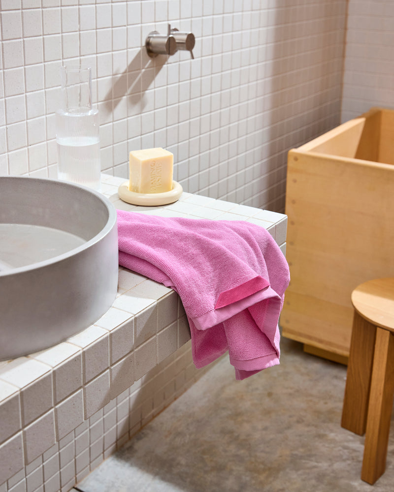 A modern bathroom with a white tiled sink, round gray basin, and a Bed Threads Bubblegum 100% Cotton Terry Hand Towel draped over the counter, with a glass of water, soap dish, wooden stool, and tub in the background.