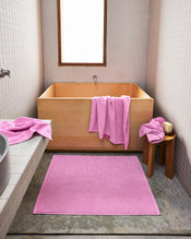 A minimalist bathroom features a light wooden tub, a pink towel on its edge, and the Bed Threads Bubblegum 100% Cotton Terry Bath Mat on the floor, with another pink towel and soap resting on a small wooden stool. - view