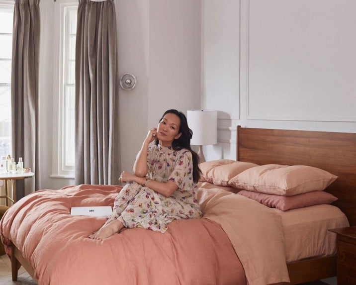 A woman in a floral dress sits on a neatly made bed with peach-colored bedding in a softly lit bedroom. There is a laptop beside her and a table with bottles near the window. Curtains frame two windows in the background.