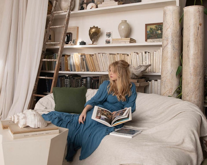 Woman in a long blue dress sits on a cream-colored couch, reading a book in a cozy room with bookshelves, decorative vases, and a wooden ladder. A green pillow and stacked books are nearby.