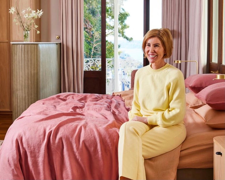 A woman in a yellow sweater and pants sits on the edge of a bed with pink bedding in a sunlit bedroom with wooden furniture and large windows showing greenery outside.