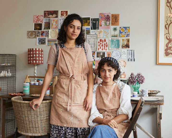 Two women wearing aprons pose in an art studio. One stands while the other sits, both surrounded by art supplies, flowers, and a wall covered with colorful art prints and cards. The atmosphere is creative and cozy.