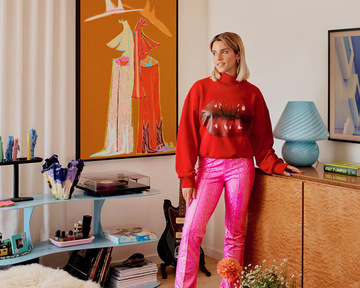 A woman in a red sweatshirt with lips printed on it and shiny pink pants stands by a wood cabinet in a colorful room decorated with art, flowers, books, a guitar, and various decorative objects.