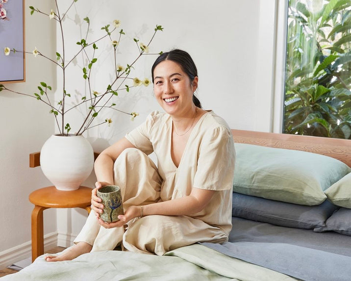 A woman in light-colored loungewear sits on a bed, smiling and holding a ceramic mug. There is a vase with branches on a small table beside her, and greenery is visible outside the window.
