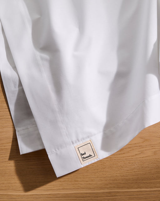 A close-up of the Crisp White 100% Organic Cotton Flat Sheet by Bed Threads shows it neatly folded on wood, featuring a small rectangular Bed Threads label sewn onto its GOTS certified fabric.