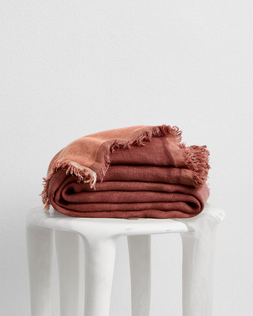 The Bed Threads Cacao & Hazelnut 100% French Flax Linen Muslin Throw with frayed edges is folded atop a white sculpted side table, set against a plain white wall.
