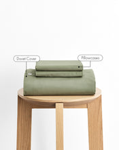 An olive green duvet cover and pillowcases, neatly folded and displayed on a light wood stool against a white background, with text labels pointing to each component. - view