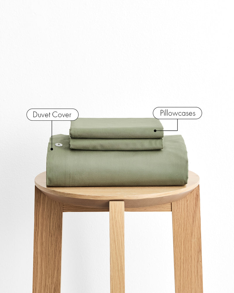 An olive green duvet cover and pillowcases, neatly folded and displayed on a light wood stool against a white background, with text labels pointing to each component.