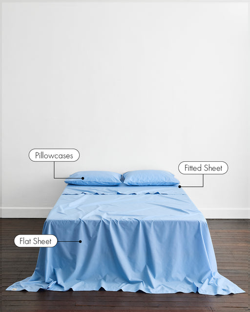 A minimalist room features a made bed with Bed Threads' Sky Organic Cotton Sheet Set (GOTS certified): pillowcases on two pillows, a fitted sheet on the mattress, and a flat sheet draped over the bed to the floor.