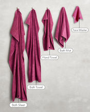 Five magenta towels from Bed Threads, including a Bath Sheet, Bath Towel, Hand Towel, Bath Mat, and a set of Berry 100% Cotton Terry Face Washers (two included), hang on hooks against a light wall. - view