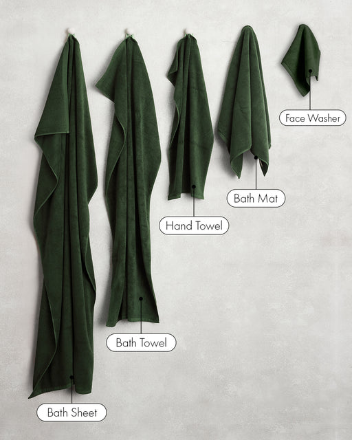 Five dark green Moss 100% Cotton Terry towels by Bed Threads—bath sheet, bath towel, hand towels, bath mat, and face washer—hang on hooks against a light wall.