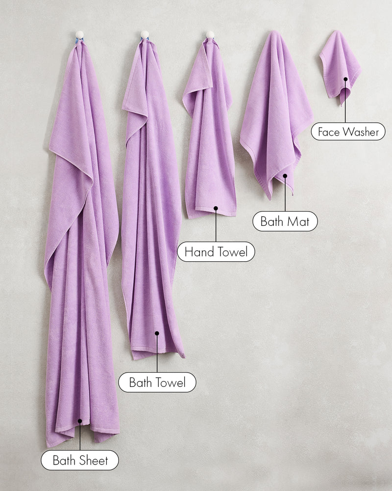 Five Bed Threads Violet 100% Cotton Terry towels in different sizes—Bath Sheet (largest), Bath Towel, Hand Towel, Bath Mat, and Face Washer (smallest)—hang on hooks against a light wall.