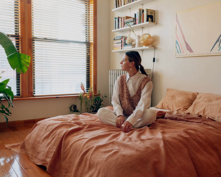 A person sits cross-legged on a peach-colored bed, looking out a sunlit window. The cozy bedroom has wooden floors, plants, wall shelves with books, and neutral-toned decor.