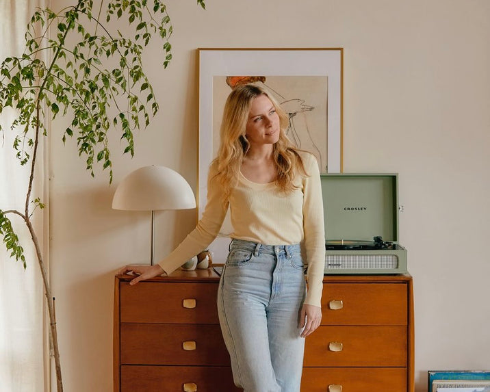 A woman with long blonde hair, wearing a yellow top and light blue jeans, stands in a cozy room by a wooden dresser with a lamp, framed art, a plant, and a green record player on top.