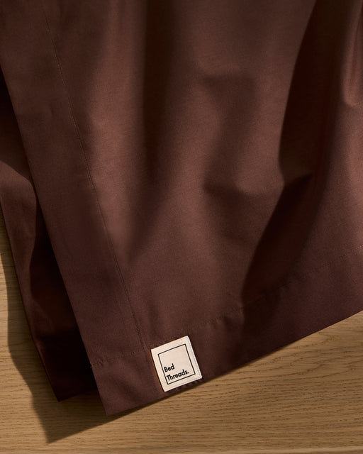 A close-up of the Espresso 100% Organic Cotton Flat Sheet by Bed Threads, GOTS certified, in dark brown with a visible hem and sewn white Bed Threads label, draped over wood.