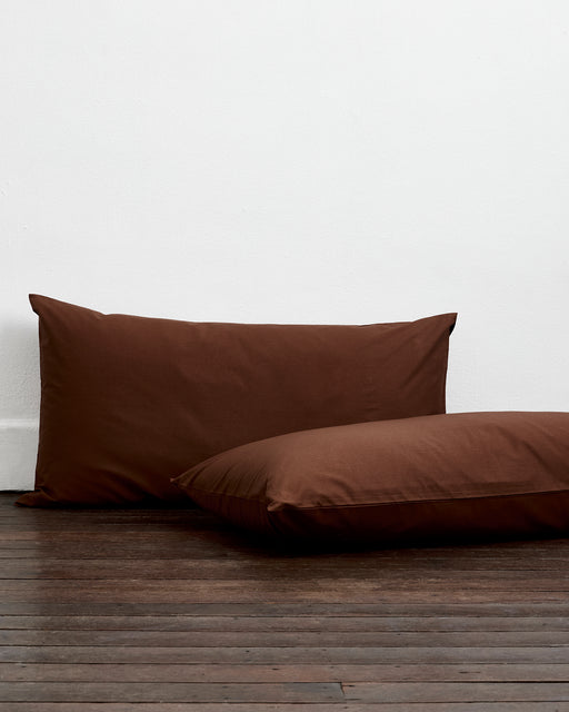 A bed with two pillows in Bed Threads Espresso Organic Cotton King Pillowcases sits against a white wall on a wooden floor, covered in brown sheets.