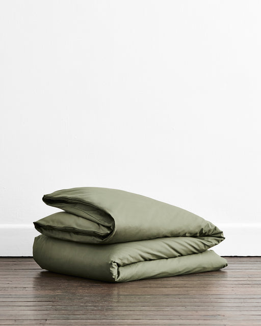 An olive green folded duvet cover, placed neatly on a dark wooden floor against a plain white wall.