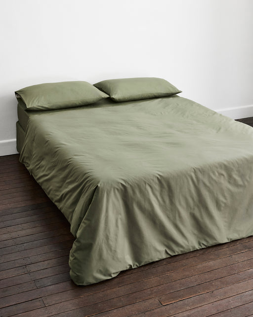 An olive green bedding set, including a duvet cover and two matching pillowcases in 100% organic cotton, neatly arranged on a bed in a minimalist room with dark wood floors and a white wall.