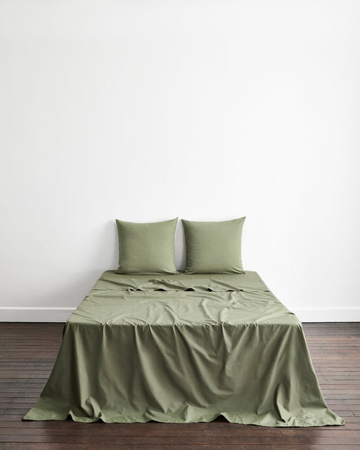 An olive green bed sheet with two matching european pillowcases on a bed, set against a plain white wall and dark wood floor.