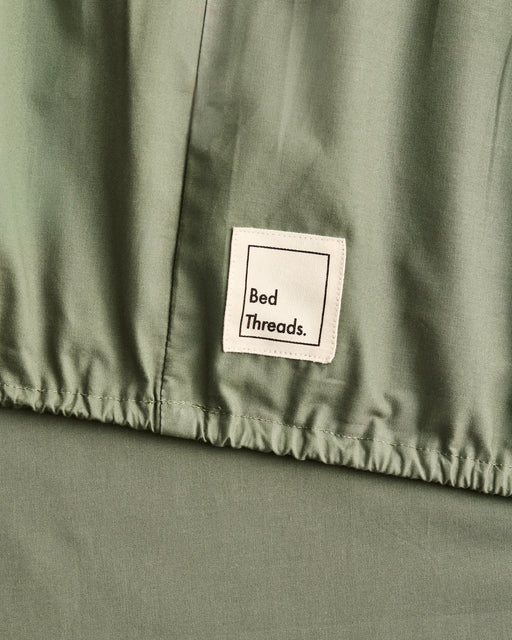 Close-up of a sage green organic cotton fitted sheet, with a white sewn-on label reading 'Bed Threads.'