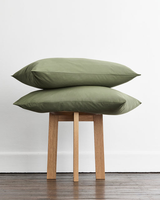 Two olive green standard pillowcases in 100% organic cotton stacked on a light wood stool against a white wall and dark wood floor.