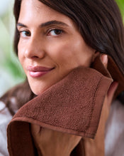 A woman smiles softly as she dries her face with a Bed Threads Espresso 100% Cotton Terry Face Washer (Set of Two), its brown, absorbent fabric complementing the calm, fresh atmosphere and providing gentle care for her skin. - view