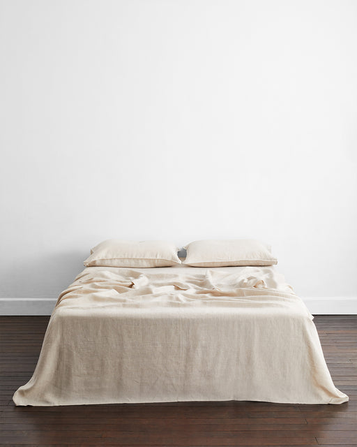 A minimalist bedroom features a low double bed dressed in Bed Threads' Oatmeal & White Stripe 100% French Flax Linen King Pillowcases, set against a plain white wall and dark wooden floors.