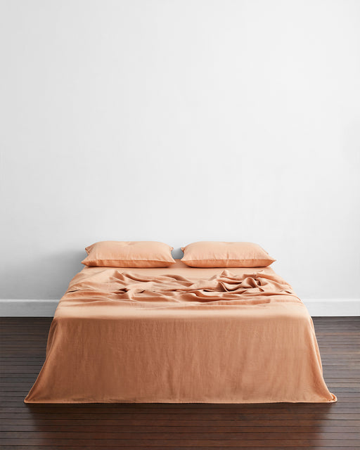 A neatly made bed with light orange sheets and Bed Threads’ Terracotta 100% French Flax Linen King Pillowcases (Set of Two) sits against a plain white wall on a dark wooden floor, offering a temperature-regulating sleep experience.