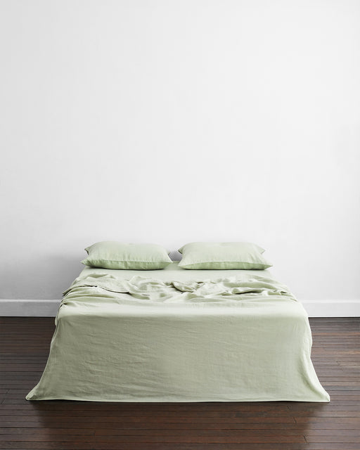 A neatly made bed with Bed Threads' Pistachio, Sage & Rosewater 100% French Flax Linen Bedding Bundle and two pillows stands against a plain white wall on a dark wooden floor in a minimalistic room.