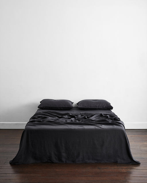 A minimalist room features a neatly made bed with Bed Threads' Charcoal 100% French Flax Linen King Pillowcases (Set of Two) against a plain white wall, offering natural temperature-regulating bedding for added comfort.