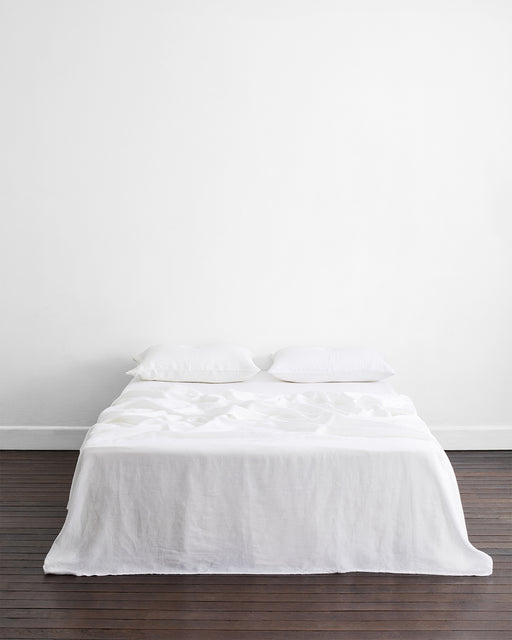 A neatly made bed with temperature-regulating bedding, white sheets, and Bed Threads’ White 100% French Flax Linen King Pillowcases (Set of Two) sits against a plain white wall on a dark wooden floor in a minimalistic room.
