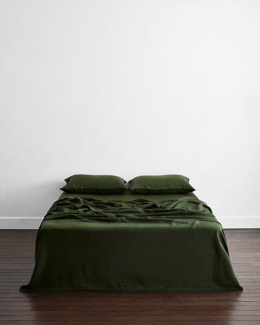 A neatly made bed features Bed Threads’ Olive 100% French Flax Linen King Pillowcases (Set of Two) in dark green, set against a plain white wall on a wooden floor in a minimal, modern bedroom.