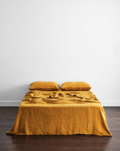 A minimalist room features a neatly made bed with mustard-yellow sheets and two Bed Threads Turmeric 100% French Flax Linen King Pillowcases, set against a plain white wall on a dark wooden floor.