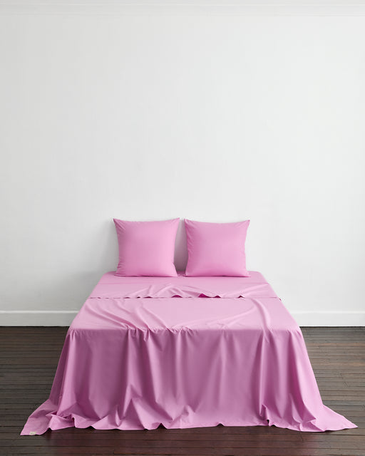 A neatly made bed with two Bed Threads Bubblegum 100% Organic Cotton European Pillowcases and a light pink sheet stands against a plain white wall on a dark wooden floor.