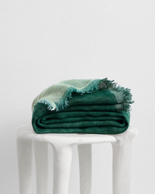 The Forest & Sage 100% French Flax Linen Muslin Throw by Bed Threads, featuring frayed edges, rests folded on a white textured stool against a plain white wall.
