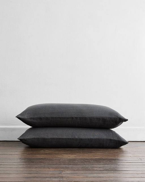 The Charcoal 100% French Flax Linen King Pillowcases (Set of Two) from Bed Threads are stacked on a wooden floor by a white wall, adding a natural, temperature-regulating accent to your space.