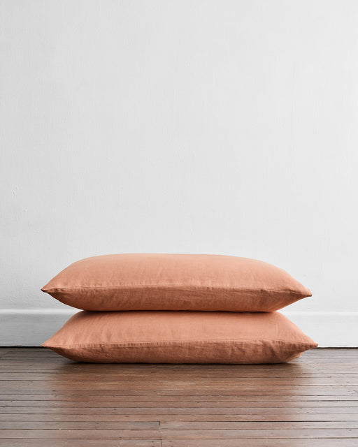 Two Hazelnut 100% French Flax Linen King Pillowcases by Bed Threads are stacked on a wooden floor against a plain white wall, providing naturally temperature-regulating comfort.