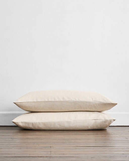 Two Oatmeal & White Stripe King Pillowcases by Bed Threads, crafted from 100% French flax linen, rest neatly stacked on a wooden floor against a white wall, offering breathable comfort in every set of two.