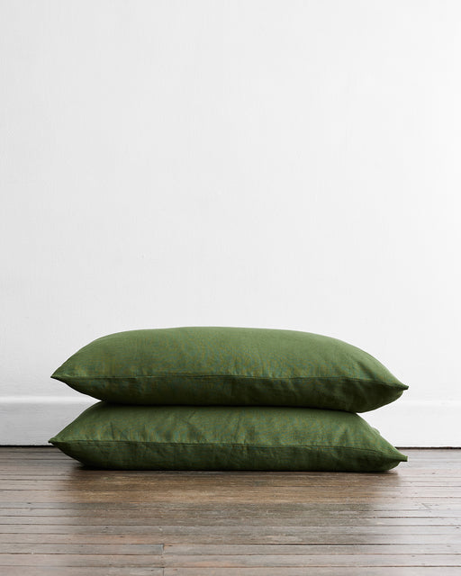 A set of two Olive 100% French Flax Linen King Pillowcases by Bed Threads is stacked on a wooden floor against a white wall, bringing a touch of breathable comfort to the space.